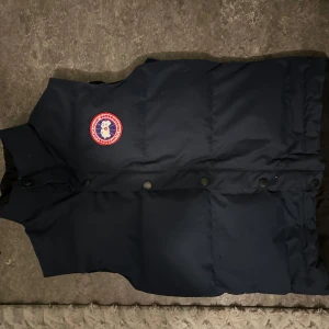 Mörkblå dunväst från Canada Goose - Säljer en snygg mörkblå dunväst från Canada Goose med ett ikoniskt märke på bröstet. Västen har knappar framtill och är perfekt för kyliga dagar. Passar både till vardags och utomhusaktiviteter. Dragkedjan på västen funkar tyvärr inte men det går att fixa för 150kr. Västen har dessutom ett litet hål där man hänger av jackan på kroken. Finns att hämta i Vårby gård. Först till kvarn som gäller!