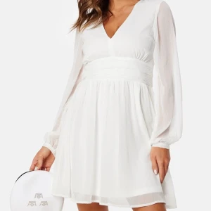 Vit klänning med v-ringning - Elegant vit klänning med v-ringning och långa ärmar. Den har en knytning i midjan och ett flödande kjolparti som ger en romantisk känsla. Perfekt för sommarens festligheter.