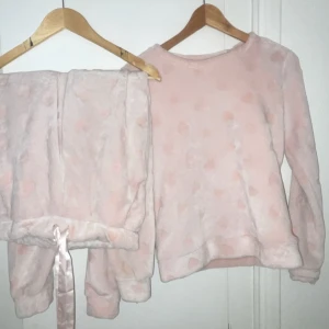 Rosa fleecepyjamas med hjärtan - Mysig rosa fleecepyjamas med hjärtmönster. Tröjan har långa ärmar och byxorna har en bekväm resår i midjan med satinband. Perfekt för kalla nätter. Byxorna har storlek xxs. Tröjan har storlek xs
