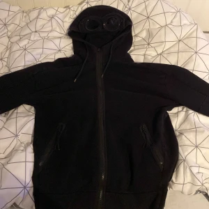 CP COMPANY Hoodie - Säljer en svart hoodie från C.P. Company med unika glasögon integrerade i huvan. Hoodien har dragkedja framtill och justerbara snören vid huvan. Perfekt för en stilren look. Hoodien är i väldigt bra skick och den ända defekten är vid fickan(bild 4).  Hoodien är i storlek XL men sitter bra på dom som har L också!