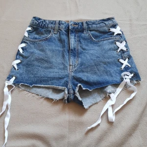 Jeansshorts med snörning från H&M Divided - Snygga blå jeansshorts från Divided med cool snörning på sidorna och fransiga kanter. Perfekta för en avslappnad sommarlook. De har en klassiska fickor och dragkedja och knapp. Använd 1 gång o har annars endast legat i låda. Skulle säga att de har medel till hög midja. 
