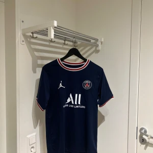 Mörkblå PSG fotbollströja - Snygg mörkblå PSG fotbollströja med korta ärmar och vit text på framsidan. Tröjan har röd, vit och blå rand vid kragen och ärmsluten. Perfekt för fotbollsfans!