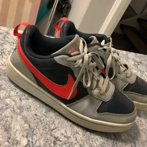 Nike sneakers i grått och rött - Snygga Nike sneakers i grått och svart med en röd swoosh. Skorna har snörning och en bekväm passform. Perfekta för en sportig look. Den har två små hål men inget som syns vid användning. Tveka inte med att ställa frågor👌
