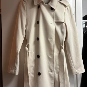 Beige trenchcoat med bälte - Snygg beige trenchcoat med knappar och bälte i midjan. Perfekt för höst och vår. Långärmad design med klassisk krage och stilrena detaljer.
