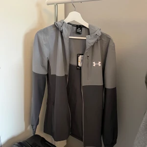 Grå vindjacka från Under Armour - Säljer en grå vindjacka från Under Armour med huva och dragkedja. Jackan har en stilren design med logotyp på bröstet och är perfekt för utomhusaktiviteter. Den är lätt och bekväm att bära. Jackan är köpt från JDsports och har aldrig hunnits användas eftersom den vart för liten och därför vill jag sälja den till ett rimligt pris. 