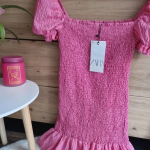 Rosa klänning från ZARA - Säljer en söt rosa klänning från ZARA med puffiga kortärmar och volangdetalj vid fållen. Klänningen har en smickrande smockad textur som ger en fin passform. Perfekt för sommarens alla tillfällen! 🌸