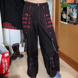 Svarta byxor med kedjedetaljer - Svarta byxor med röda detaljer och kedjor. Byxorna har en loose passform och är ganska tunna i materialet. Passar personer som har en alternativ/punkig stil. Byxorna är knappt användna och har bara en defekt. En strapdetalj längst ner på högra byxbenet har sömen gått upp lite grann