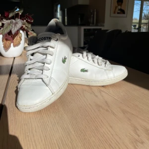 Vita sneakers från Lacoste - Snygga vita sneakers från Lacoste med klassisk design och snörning. Skorna har det ikoniska krokodilmärket på sidan och en bekväm passform. Perfekta för en stilren look. Det finns några slitningar men inga större (se bilder) jätte fina skor att ha på sig.