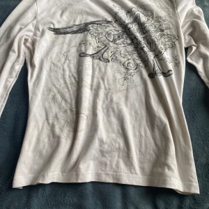 Vit långärmad tröja med tryck y2k - Säljer en vit långärmad tröja med ett detaljerat svart tryck på framsidan. Tröjan har en normal passform och är perfekt för en avslappnad stil. Materialet känns mjukt och bekvämt mot huden.