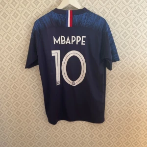 Mbappe fotbollströja 2018  - Säljer en blå fotbollströja från Nike med nummer 10 och namn på ryggen. Tröjan har korta ärmar och ett stilrent mönster i blått och vitt. Perfekt för fotbollsfans!