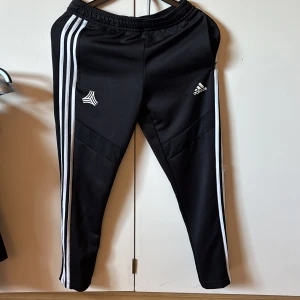 Svarta träningsbyxor från Adidas - Säljer ett par svarta träningsbyxor från Adidas med klassiska vita ränder längs sidorna. Byxorna har en elastisk midja och är perfekta för sport och träning. Adidas-loggan är tryckt på framsidan.