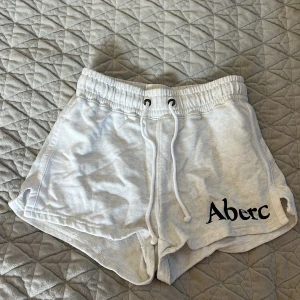 Vita shorts från Abercrombie & Fitch - Säljer ett par vita shorts från Abercrombie & Fitch med elastisk midja och snörning. Perfekta för en avslappnad stil. Loggan är tryckt i svart på sidan.