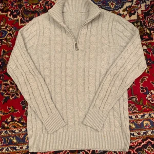 Grå stickad half zip - Säljer en mysig grå stickad tröja med dragkedja och hög krage. Tröjan har ett klassiskt kabelstickat mönster och långa ärmar. Perfekt för kyliga dagar!