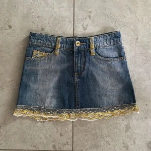 Jeanskjol med spetsdetaljer - Så fin jeanskjol i blått med snygga spetsdetaljer i gult och vitt längs fållen. Kjolen har klassiska femfickor och bälteshällor, perfekt för en avslappnad stil. Midjemått tvärsöver: 31 cm. 