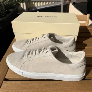 Beige sneakers från Axel Arigato - Snygga beige sneakers från Axel Arigato i mocka med vit sula. Skorna har en stilren design med snörning och diskret logga i guld på sidan. Perfekta för en avslappnad och trendig look.