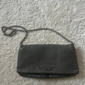 Grå axelväska från Zadig & Voltaire - Snygg grå axelväska i skinn från Zadig & Voltaire med kedjerem och dragkedja. Säljer billigt pga skicket och att jag inte har något kvitto🥰