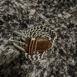 Flätat bälte med läderdetaljer - Snyggt flätat bälte  i svart och beige med bruna läderdetaljer. Perfekt för att ge en stilren touch till din outfit. Spännet är i silverfärgad metall.