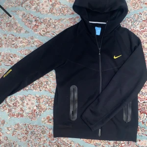 Svart Nike Nocta Tech - Säljer en svart Nike tech från Nocta och text på ärmen. Den har dragkedja framtill och två fickor med dragkedjor. Perfekt för en sportig look eller avslappnad stil.