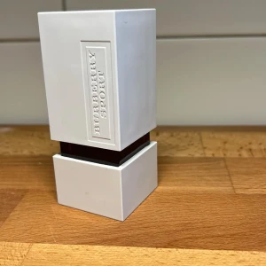 Burberry Sport Parfym - Säljer en stilren Burberry Sport parfym i en vit, fyrkantig flaska med en modern design. Flaskan har en präglad logotyp på framsidan och en kontrasterande mörk detalj i mitten. Perfekt för den som gillar en sportig och fräsch doft.