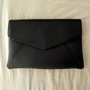 Svart kuvertväska med kedja - Elegant svart kuvertväska med en stilren design och en guldfärgad kedja som axelrem. Väskan har en dekorativ tofsdetalj på sidan. Hade den till balen med kedjan och tofsen instoppad 🤍