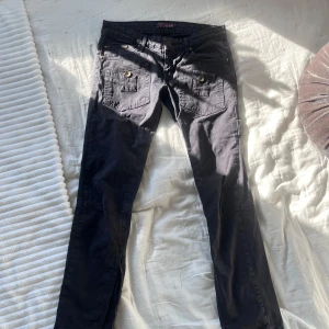 Svarta lågmidjade jeans med knappar - 💛Snygga svarta byxor med flera fickor och coola detaljer som dragkedjor vid bensluten. Byxorna har knappar både fram och bak, vilket ger en unik look. Perfekta för en avslappnad stil. De är mjuka att röra på! 💛            •Midjemåttet: 37,5 cm rakt över och 75 cm runt hela.           •Innerbenslängd: 72cm 