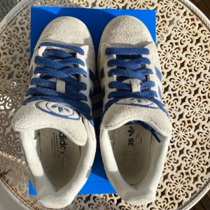 Adidas Campus sneakers i beige/vit och blå - Snygga Adidas Campus sneakers i beige mocka med blå detaljer och snörning. Det medföljer samma kartong som jag fick när jag köpte de, även ett par vita skosnören följer med! Använt endast lite nu i vår och säljer för att jag vill köpa andra skor, tyckte inte jag passade i de. Skriv gärna innan du vill köpa! Pris kan diskuteras!!