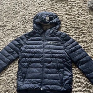 Mörkblå dunjacka från Sail Racing - Säljer en mörkblå dunjacka från Sail Racing med huva och dragkedja. Jackan har en quiltad design och är perfekt för vår väder, inte för varm och inte för kall. Insidan är ljusgrå och jackan har praktiska fickor med dragkedjor.