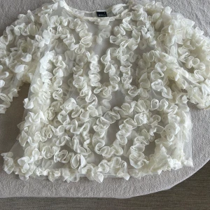 Vit blus med volanger - Säljer en elegant vit blus med volanger över hela ytan. Blusen har en lös passform och trekvartsärmar, perfekt för en stilfull look. Passar bra till både jeans och kjol för en chic outfit.