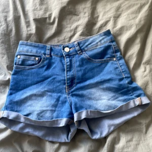 Blå jeansshorts från Noisy May - Snygga blå jeansshorts från Noisy May med klassisk femficksdesign och uppvikta benslut. Perfekta för en avslappnad sommarlook. De har en knapp och dragkedja framtill för enkel stängning.