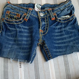 Blå jeansshorts från True Religion - Säljer ett par coola blå jeansshorts från True Religion med snygga kontrastsömmar och slitna detaljer. Shortsen har klassisk femficksdesign och en knappgylf. Perfekta för en avslappnad stil!