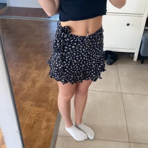 Svart blommig kjol med volang - Säljer en söt svart kortkjol med vita blommor och volangdetaljer. Kjolen har en omlott-design med knytning i midjan, vilket ger en fin passform. De har även som shorts under vilket gör att kjolen ej blåser upp. 