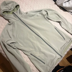 Ljusgrön fleecejacka från Houdini - Säljer en stilren ljusgrön fleecejacka från Houdini med huva och hel dragkedja. Jackan har två praktiska fickor med dragkedja och är perfekt för lager-på-lager. Mjuk och skön, passar dig som gillar outdoor och chill vibes.