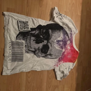 Vit t-shirt Concrete Humanity/Philipp Plein - Vit t-shirt från Concrete Humanity med stor svart dödskalle framtill, färgstänk i lila, rött och gult samt tryck på ärmarna. Baksidan har röd graffiti-text och smiley. Skön passform och cool streetstil, perfekt för dig som gillar unika prints.