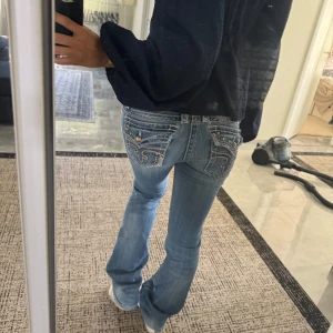 Blå bootcut jeans från Rock Revival - Snygga ljusblå bootcut jeans från Rock Revival med coola broderade bakfickor och silvriga detaljer. Jeansen har låg midja och klassisk femficksdesign. Materialet är jeans med stretch för skön passform. Perfekta för dig som gillar detaljer och en unik look.// Kolla bilder för skick och mått!!!! Innerbenslängd: 84cm