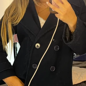 Mörkblå skepparkavaj från Design by Si - Säljer en snygg mörkblå skepparkavaj från Design by Si. Jackan har dubbelknäppning med stora knappar och bred krage. Tillverkad i ullblandning, perfekt för kyliga dagar. Klassisk och tidlös modell som är enkel att styla till olika outfits.