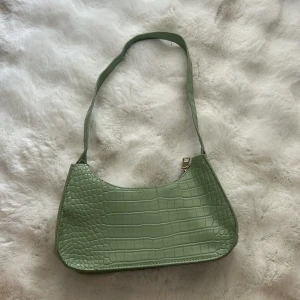 Grön axelväska med krokoprägling - Snygg ljusgrön axelväska med krokodilpräglat mönster och dragkedja upptill. Väskan har en enkel, stilren design och är tillverkad i syntetmaterial. Perfekt storlek för det viktigaste och lätt att bära över axeln.