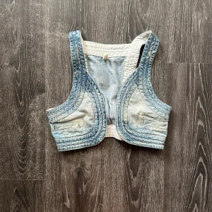 Ljusblå croppad jeansväst - Säljer en croppad ljusblå jeansväst med coola sömdetaljer och justerbart spänne i ryggen. Västen har en sportig form, vita prickiga insidor och fejkfickor framtill. Perfekt att styla över en t-shirt eller klänning för en trendig look. 36/34 ungefär 