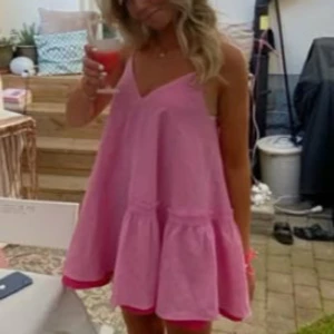 Rosa volangklänning med v-ringning - Supersöt rosa kortklänning med volangkant och v-ringad hals. Klänningen är ärmlös med tunna axelband och har en luftig, loose passform – perfekt för varma dagar. Enkel och trendig design som ger en avslappnad men ändå chic vibe.