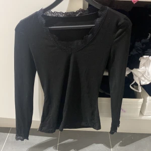 Svart långärmad topp med spets - Svart långärmad topp med v-ringning och fina spetsdetaljer vid halsringningen och ärmsluten. Snygg och enkel modell som passar perfekt till jeans eller kjol. Mjuk och stretchig känsla för en bekväm passform.