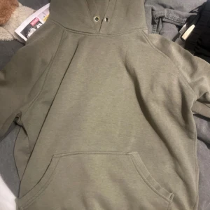 Olivgrön hoodie med magficka - Olivgrön hoodie med klassisk magficka och huva. Tröjan har långa ärmar och är tillverkad i mjukt bomullsmaterial. Perfekt för chill dagar eller när du vill ha en enkel och stilren look.