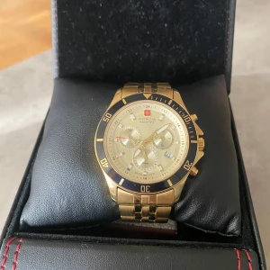 Swiss Military Hanowa guldig klocka - Snygg guldig klocka från Swiss Military Hanowa med robust länkarmband i metall. Urtavlan är ljus med flera små ur och svarta detaljer runt boetten. Klockan har kronograf och datumvisning, kommer även i originalbox med extra länkar. Klockans batteri är dessutom ny bytt. 