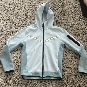  Nike tech fleece ljusblå  - Nike tech  i ljusblått med hel dragkedja och huva. Jackan har vita detaljer, svarta kanter och en praktisk ficka med dragkedja på ärmen. Perfekt för dig som gillar sportig stil och vill ha något lätt och snyggt till vardags.