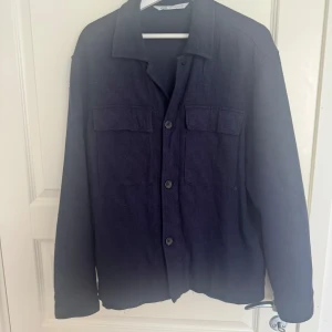 Zara overshirt - Snygg mörkblå overshirt från Zara med klassisk krage och två stora bröstfickor. Jackan har knäppning framtill med knappar och är tillverkad i ett mjukt tyg som känns skönt mot huden. Perfekt att slänga över en hoodie eller t-shirt för en chill look.