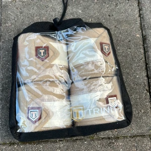 Beige lindor från Trinity - Säljer ett par snygga beigea lindor från Trinity. De har en stilren designpå och är gjorda i mjukt fleece-liknande material med detaljer i satin. Perfekta för dig som vill ha både stil och funktion i stallet.