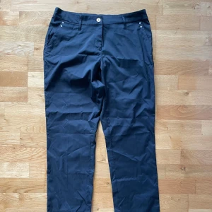 Svarta golfbyxor från Abacus, stl 40 - Svarta golfbyxor från Abacus i klassisk rak modell med fickor fram och bak. Byxorna har bälteshällor och diskreta silvriga nitar vid fickorna. Perfekta för en clean och stilren look.