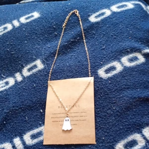 Guldigt halsband med spökhänge - Snyggt halsband med tunn guldfärgad kedja och ett vitt spökhänge med svarta ögon. Perfekt accessoar för dig som gillar unika och lekfulla smycken. Kedjan är enkel och klassisk medan hänget ger en cool twist.