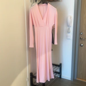 Ljusrosa långklänning med v-ringning - Säljer en elegant ljusrosa långklänning med djup v-ringning och långa ärmar. Klänningen har en figurnära passform och är tillverkad i ett mjukt, stretchigt material som faller snyggt längs kroppen. Perfekt för dig som vill ha en stilren och feminin look.
