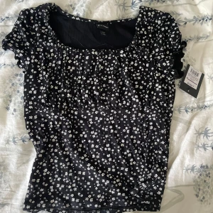 Svart blommig topp från Art Class - Supersöt svart topp från Art Class med små vita blommor över hela tyget. Toppen har korta ärmar med volangdetaljer och en rundad halsringning. Materialet är ribbat och stretchigt, vilket gör den både bekväm och snygg. Perfekt för dig som gillar en feminin och trendig stil. Aldrig använd.     