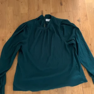 Mörkgrön blus från Holly & Whyte - Snygg mörkgrön blus från Holly & Whyte med rund halsringning och lätt puffade axlar. Långa ärmar och knapp i nacken ger en stilren look. Perfekt att matcha med jeans eller kjol för en trendig outfit.