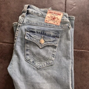 True Religion Joey Low Rise Flare Jeans - Säljer ett par ljusblå True Religion Joey jeans med låg midja och utsvängda ben. Klassiska bakfickor med lock och tydliga sömmar, silverfärgade knappar och detaljer. Jeansen är i mjukt denim och har en snygg tvättad look.